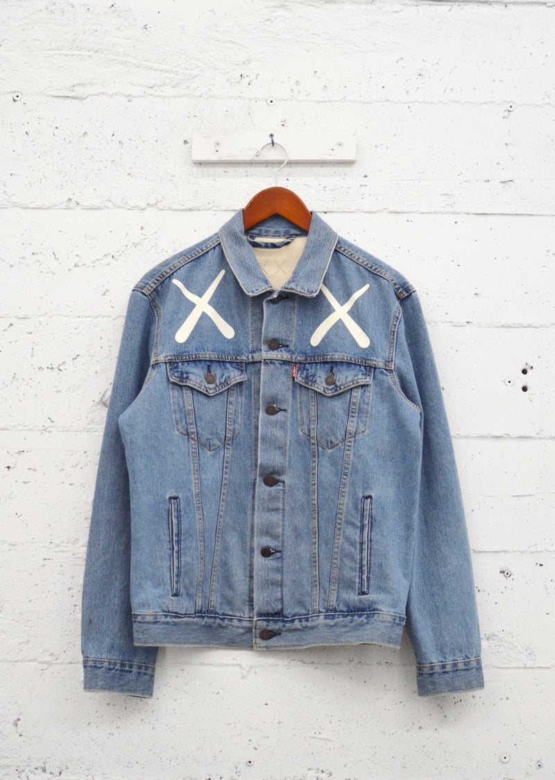 In-House custom denim trucker jacket with bold XX graphic print hanging against a white-painted brick wall.