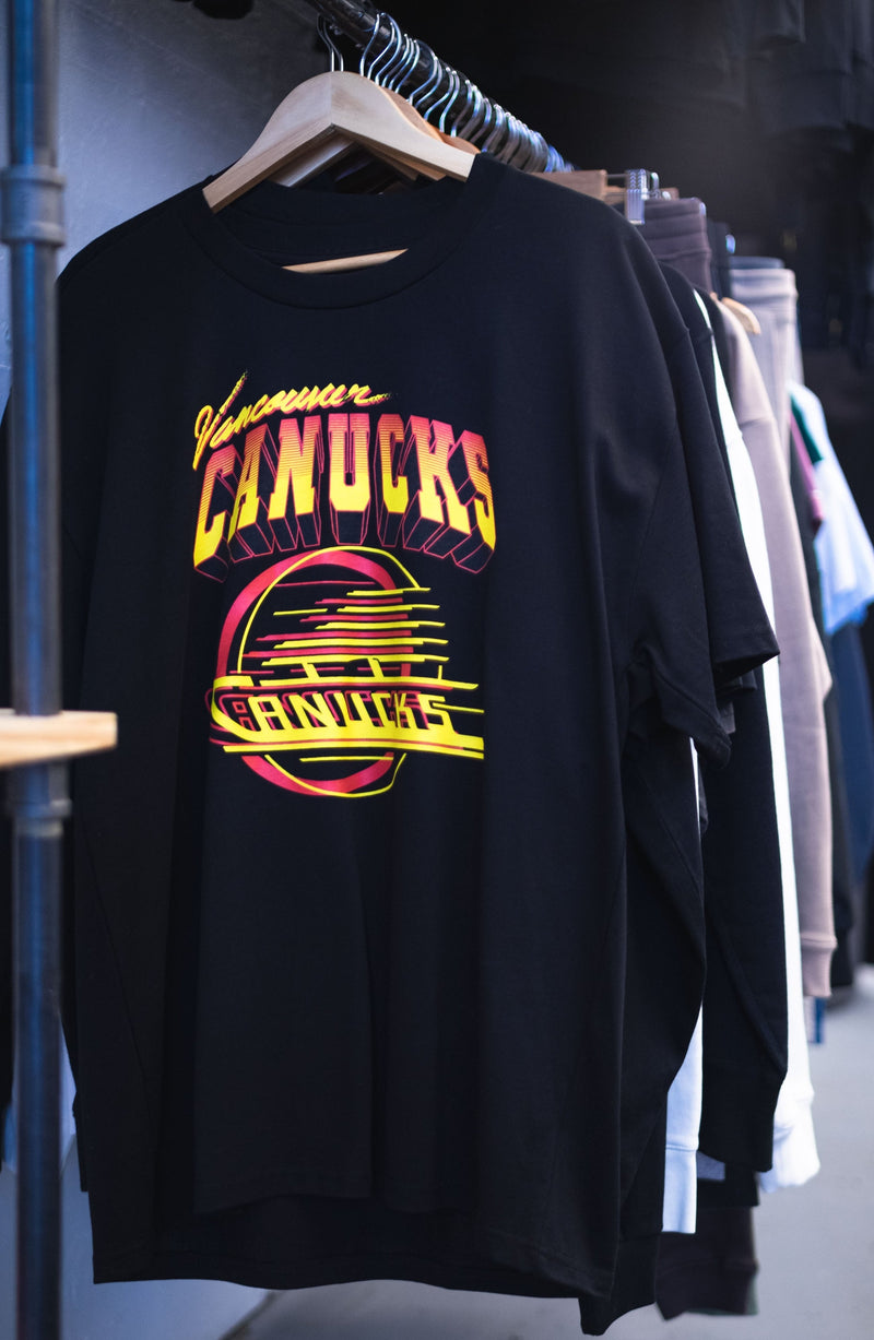 A black t-shirt with a bright yellow and red Vancouver Canucks logo hanging on a rack created by In House.