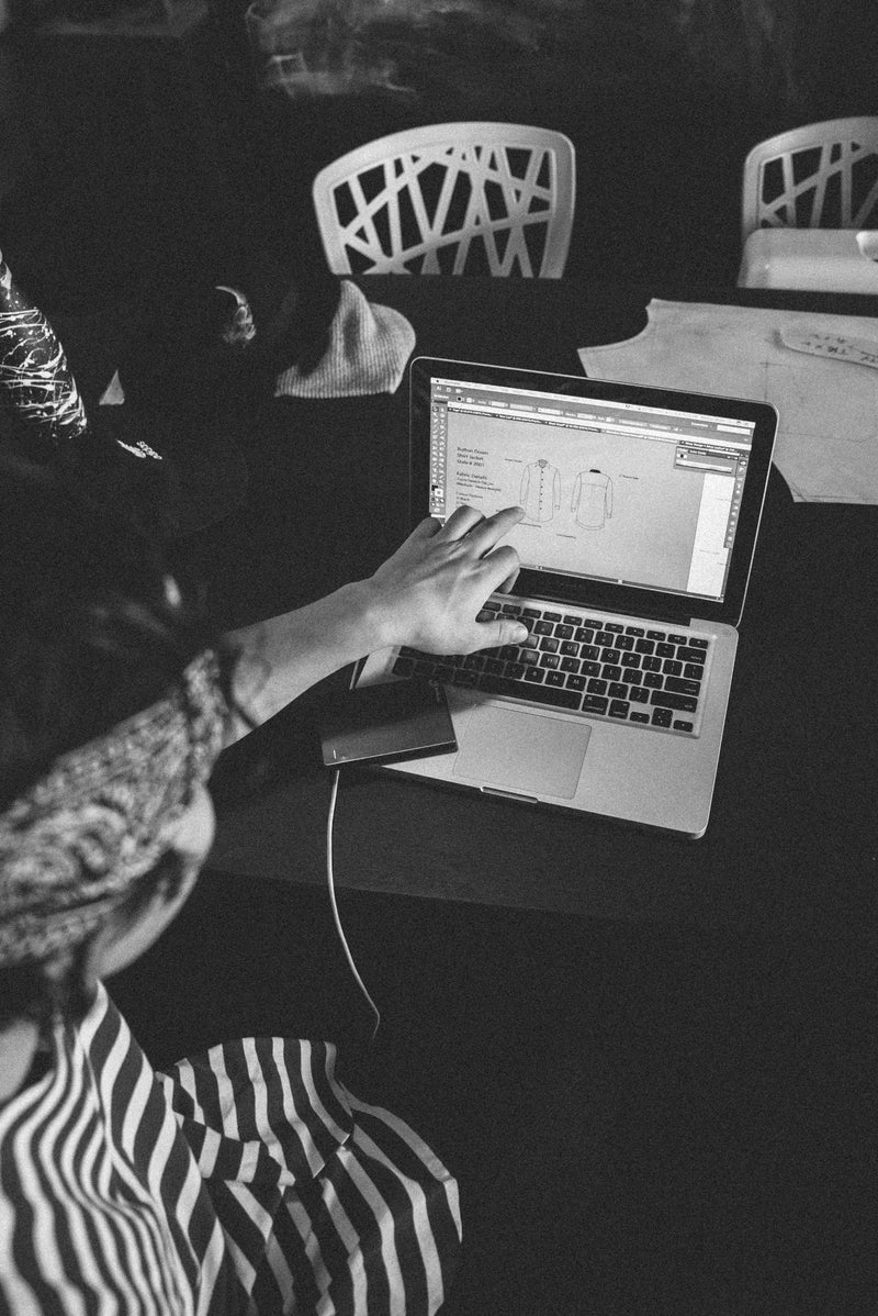 Black and white photo of a person pointing at a garment spec on a laptop screen.
